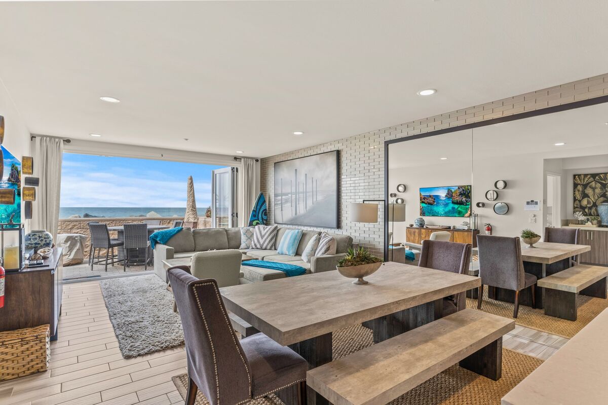 An open living area showcases modern furnishings, including a large sectional sofa and a dining table with bench seating. Sliding glass doors lead to a private patio with ocean views, enhancing the space with natural light and a connection to the outdoors.
