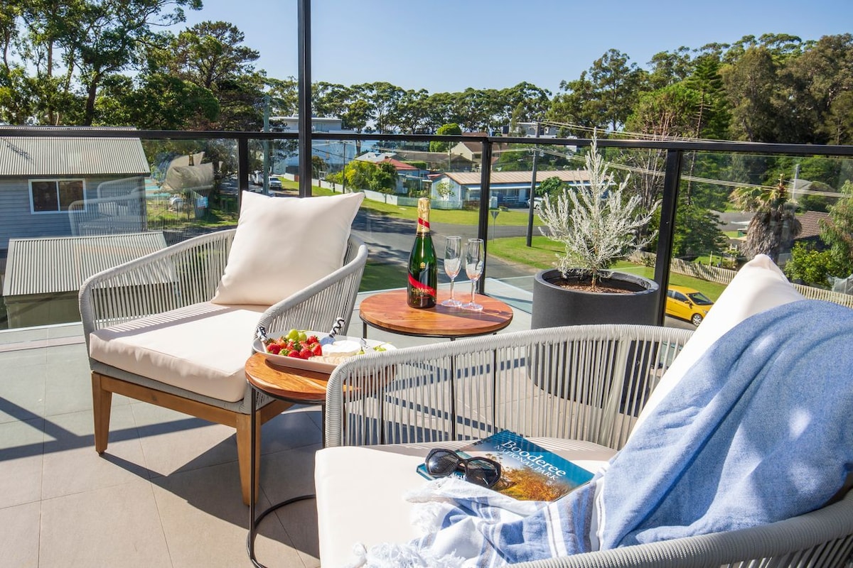 A terrace features two comfortable lounge chairs paired with a small side table. A bottle of champagne and two glasses sit on the table, accompanied by a plate of fresh strawberries. Green trees and residential views are visible in the background.