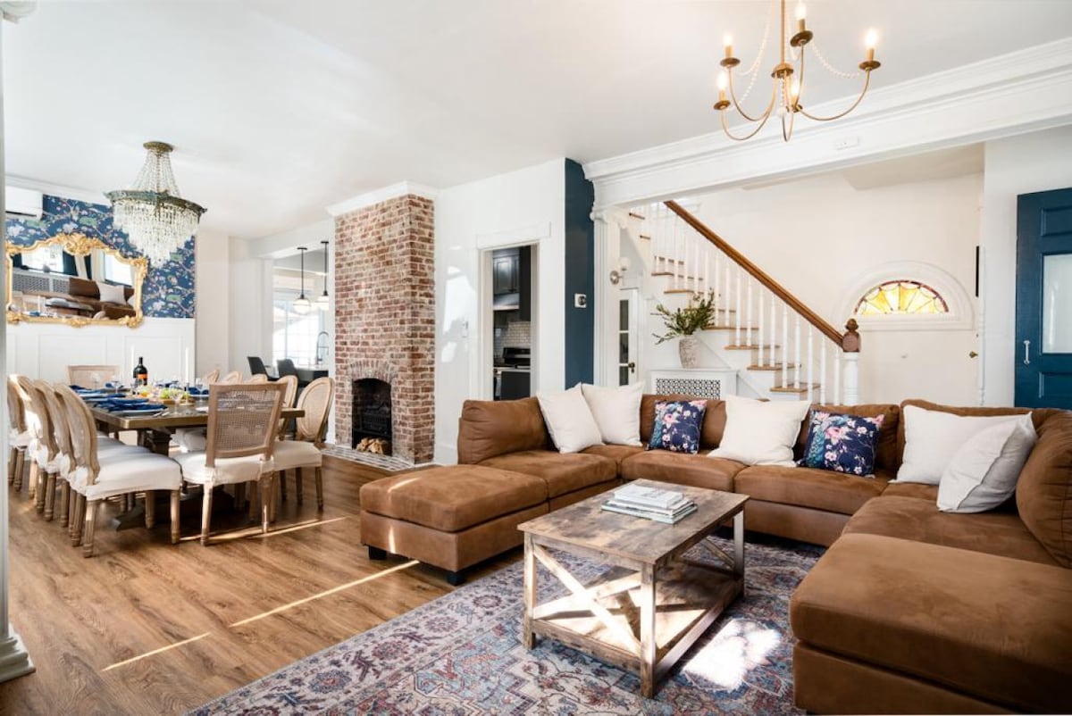 A spacious living area features a sectional sofa adorned with decorative pillows, a rustic coffee table, and wooden flooring. Natural light filters through large windows, highlighting the brick fireplace and a dining area with elegant chairs. A chandelier hangs from the ceiling, adding a distinctive touch.