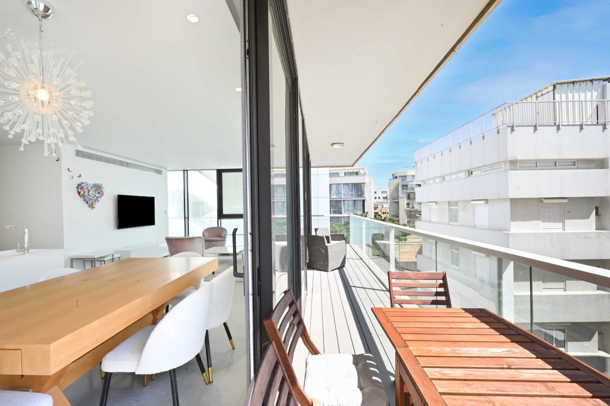 A spacious balcony presents a blend of relaxation and dining areas. A long wooden table, accompanied by white chairs, sits alongside a smaller wooden table with matching chairs. Expansive glass doors invite natural light, offering views of the surrounding buildings and clear blue skies.