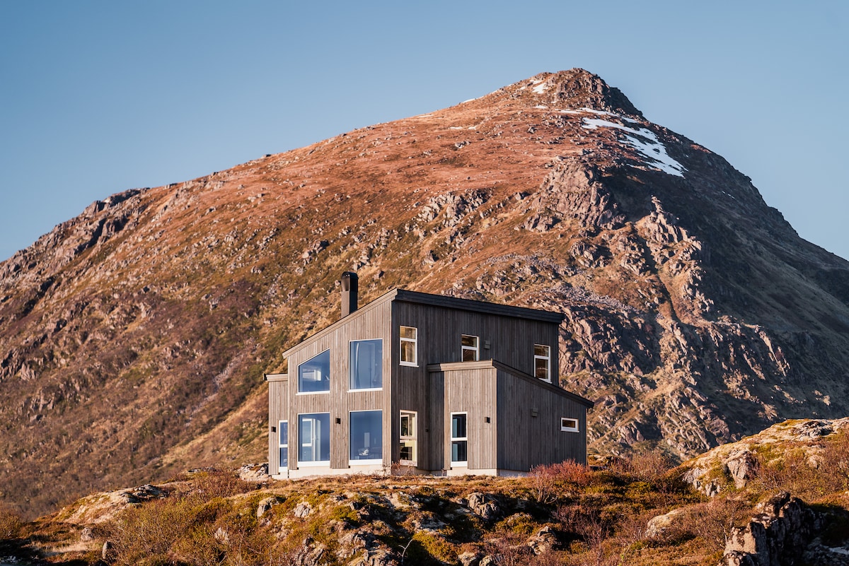 A modern cabin is positioned on a rocky landscape, surrounded by mountains. Large windows allow for ample natural light and showcase the stunning scenery. The structure features a sleek design with wood siding, reflecting a harmonious connection with the natural environment.