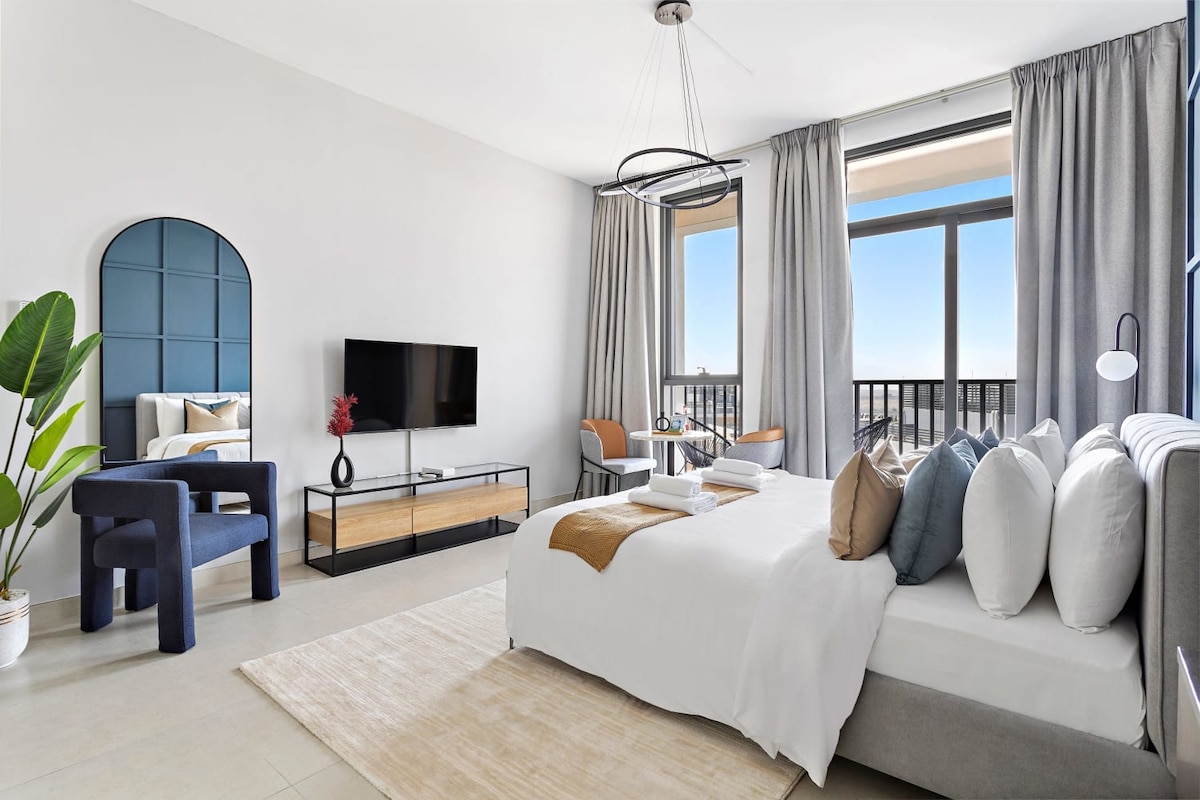 A modern bedroom is highlighted by a spacious bed adorned with decorative cushions. A sleek television is mounted on the wall opposite the bed. Two windows provide natural light, complemented by soft curtains. A cozy seating area with stylish chairs and a small table is visible.