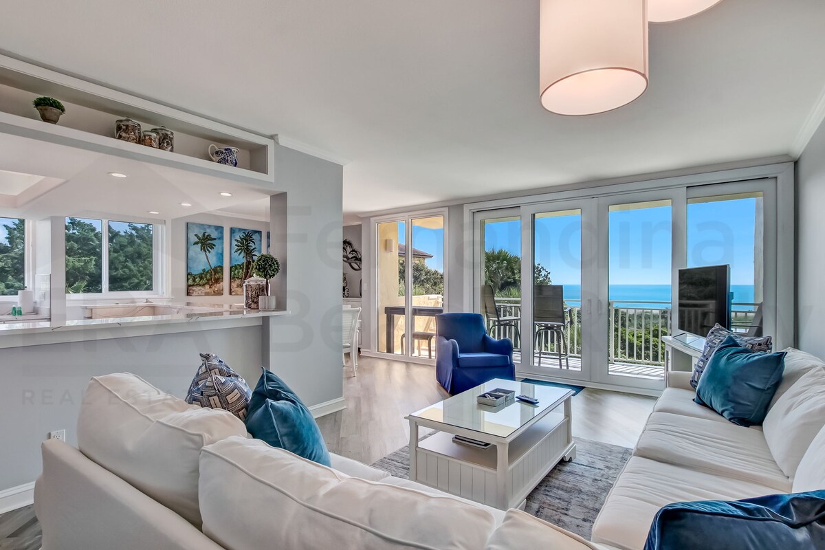A light-filled living area is showcased, featuring a comfortable white sofa with decorative pillows. Large glass doors open to reveal ocean views, while a blue armchair adds a pop of color. The open layout connects to a dining space and kitchen, emphasizing social interaction.