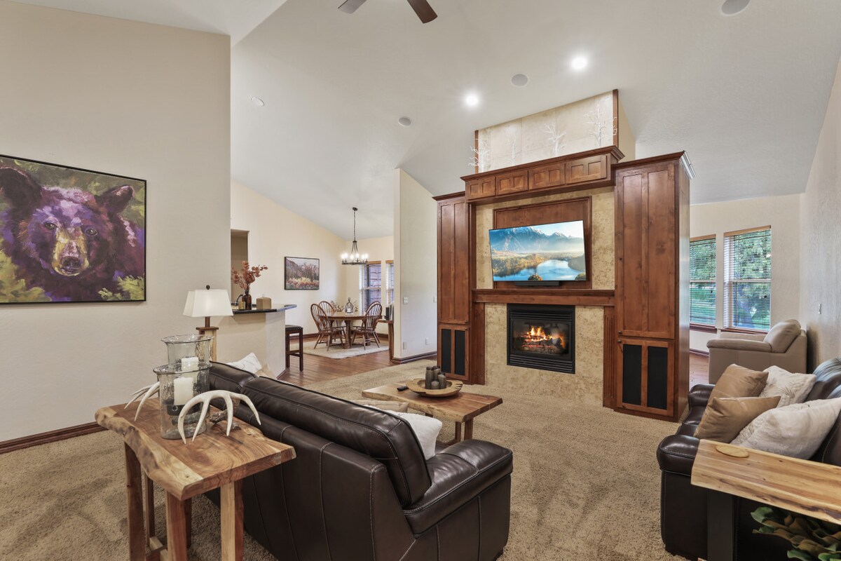 A spacious living area features high ceilings and natural light from large windows. A stone fireplace serves as a focal point, complemented by wooden cabinetry. Comfortable seating is arranged around a coffee table, with wildlife art adding character to the walls.
