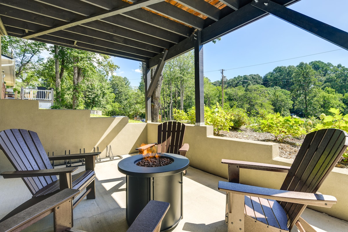 A covered patio area features a circular fire pit surrounded by four wooden Adirondack chairs. Green foliage and trees provide a natural backdrop, with clear blue skies visible overhead.