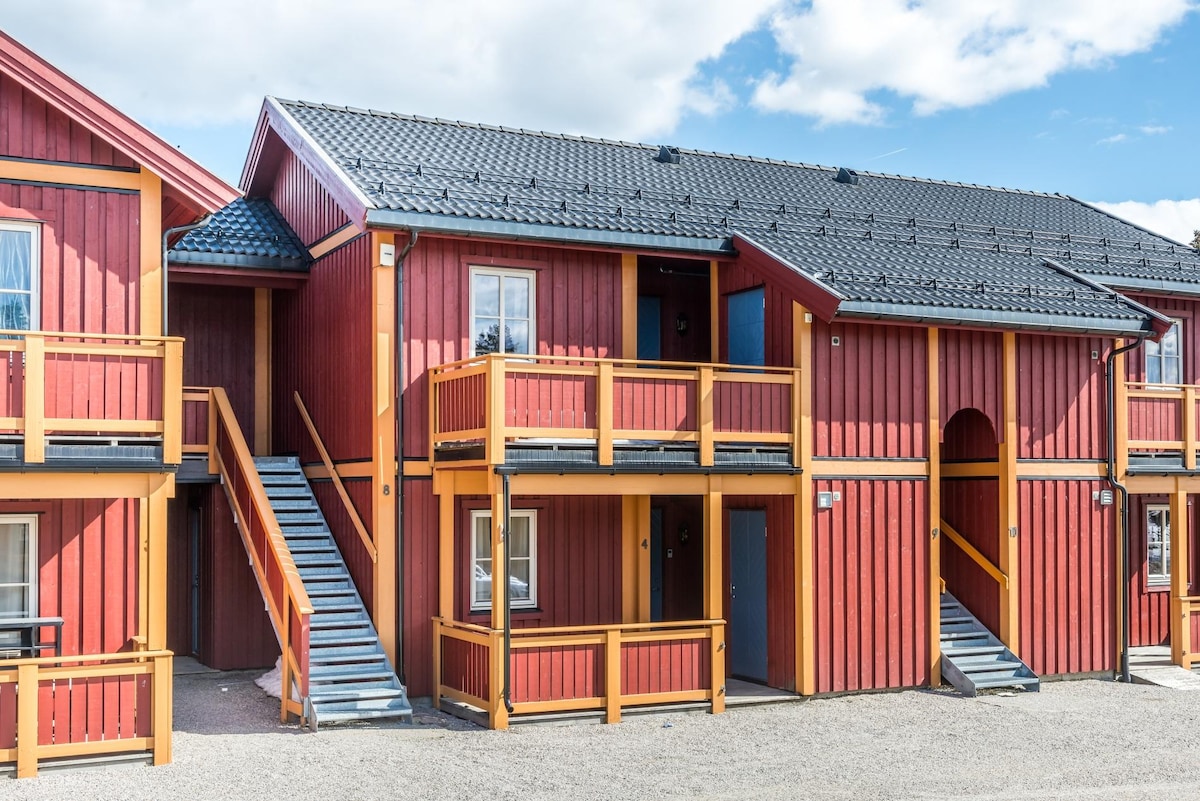 A vibrant red structure features two floors with multiple entrances, each accompanied by wooden railings. Sturdy staircases lead to the upper level, providing access to the balcony areas. The surrounding gravel area enhances the building's rustic charm against a blue sky with scattered clouds.
