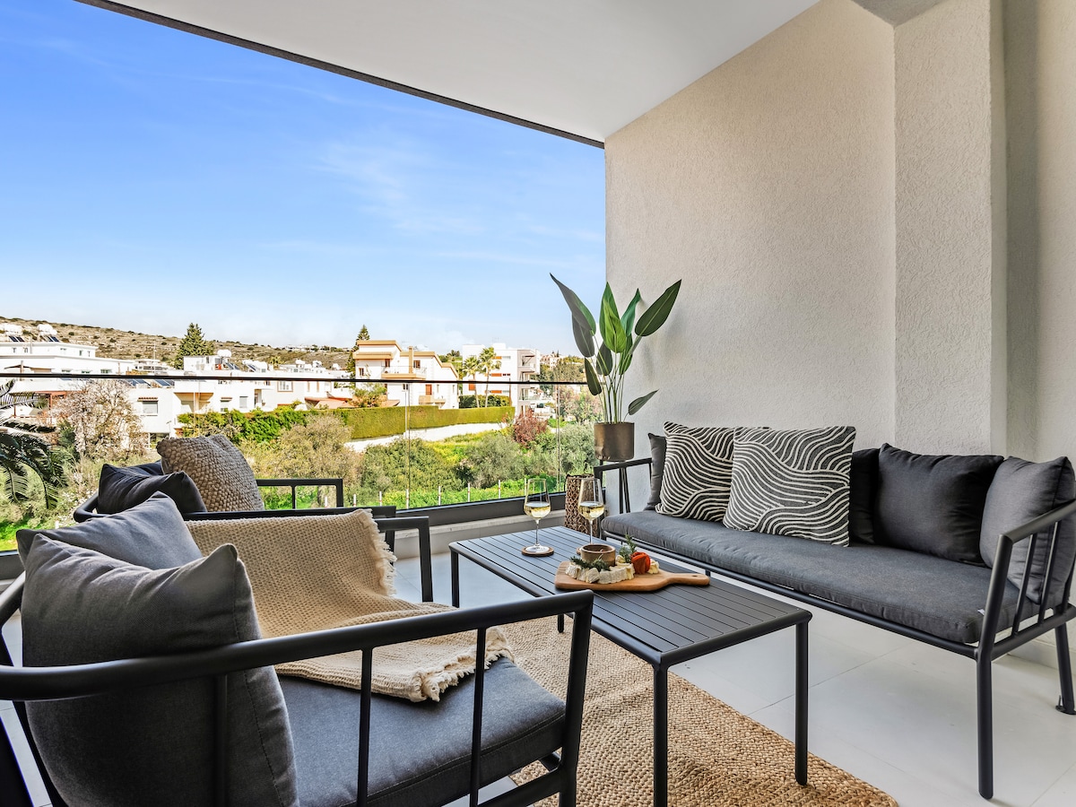 A spacious balcony features stylish seating arrangements, including a two-seater and a loveseat. A table, set with refreshments, enhances the inviting atmosphere. Lush greenery and a distant view of the neighborhood provide a serene backdrop, complemented by the bright blue sky overhead.