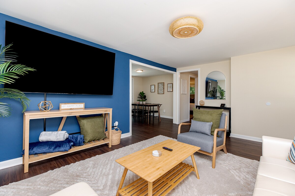 A bright living area features a large flat-screen TV mounted on a blue accent wall. A light wooden coffee table sits on a cozy area rug, surrounded by two armchairs. The space is complemented by a decorative plant and an inviting atmosphere.