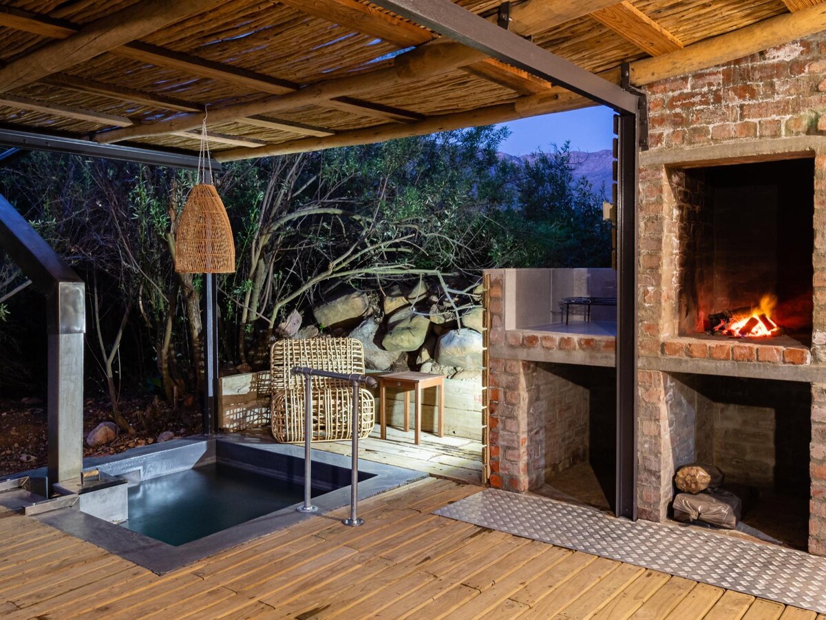 An outdoor space features a wood-fired heated tub beside a shaded covered patio. A stone-built braai stands ready for grilling, with fire visible inside. Two wicker chairs are positioned nearby, set against a natural backdrop of greenery and rocks.