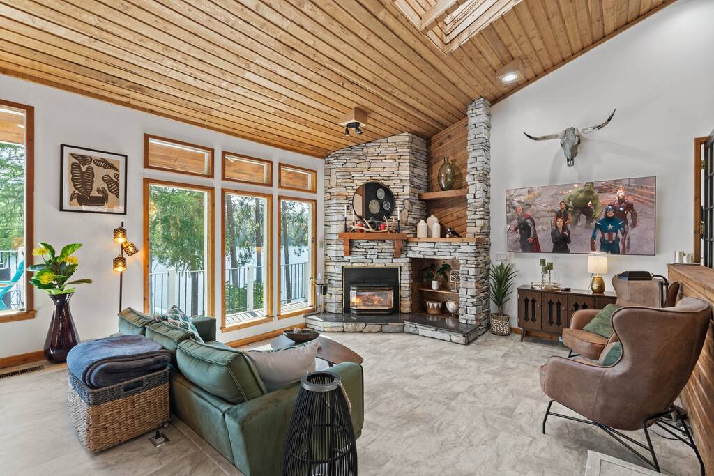 The bright living space features a stone fireplace with a wooden mantel and a large wall-mounted TV. Natural light floods through multiple windows, highlighting the earthy tones of the furnishings, including a sectional sofa and a cozy chair. A decorative cow skull and wall art add unique touches.