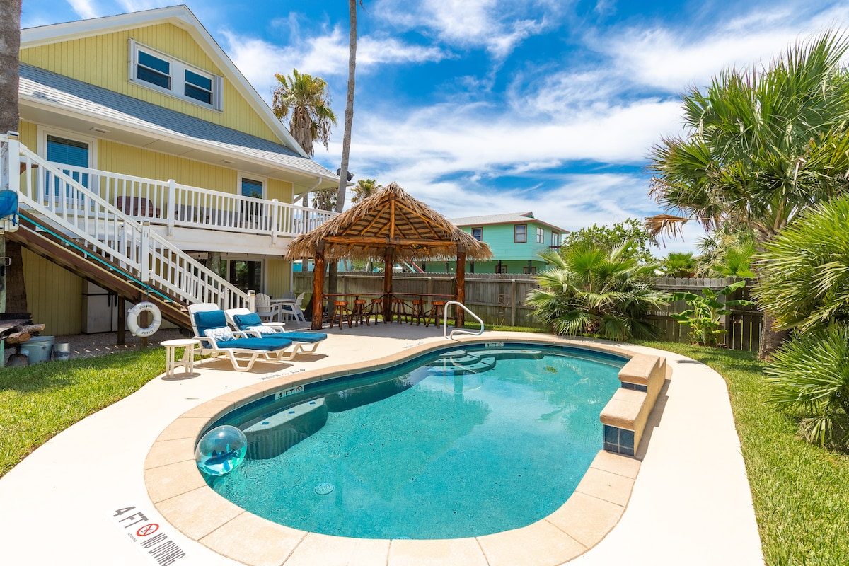 A private pool is framed by lush landscaping, featuring a shaded palapa with barstool seating nearby. Comfortable lounge chairs are positioned by the water’s edge, while a two-story home stands in the background under a clear blue sky.