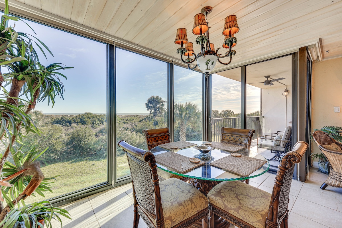 A dining area is presented with a glass table surrounded by wicker chairs, complemented by a chandelier. Floor-to-ceiling windows offer expansive views of the natural surroundings, allowing ample light to fill the space.
