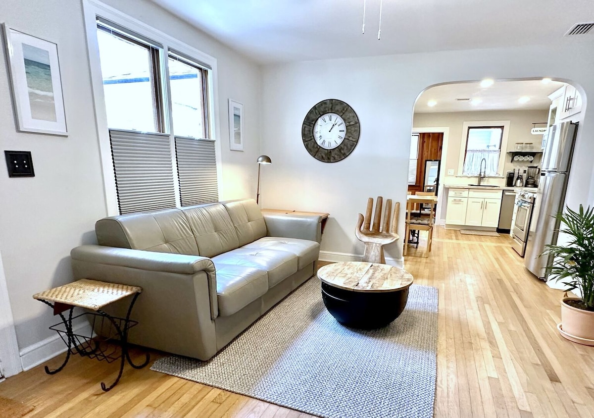 A light-filled living area features a comfortable leather sofa and a circular wooden coffee table atop a textured rug. A pair of windows offers natural light, while a wall clock adds a functional element. A dining table with wooden chairs is visible leading into the kitchen area.