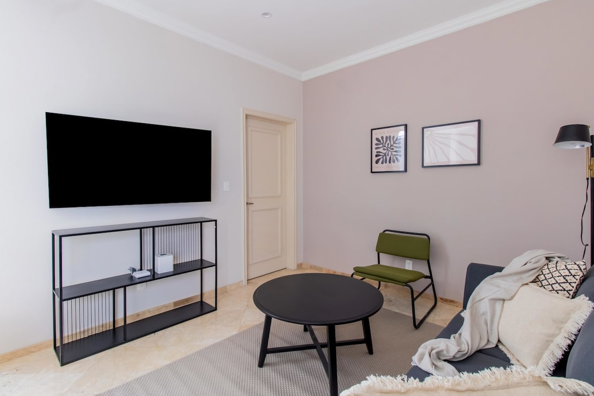A modern living area features a sleek black TV mounted on the wall and a minimalist black coffee table centered on a light-colored rug. A green accent chair provides additional seating, complemented by soft wall art framed in black. A neutral tone decorates the overall space.