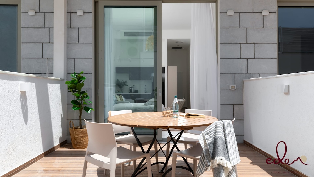 An outdoor seating area features a round wooden table surrounded by four white chairs. A small green plant decorates the corner, and a gray blanket is draped over one chair. Natural light enters through large glass doors, leading to a bright interior space.
