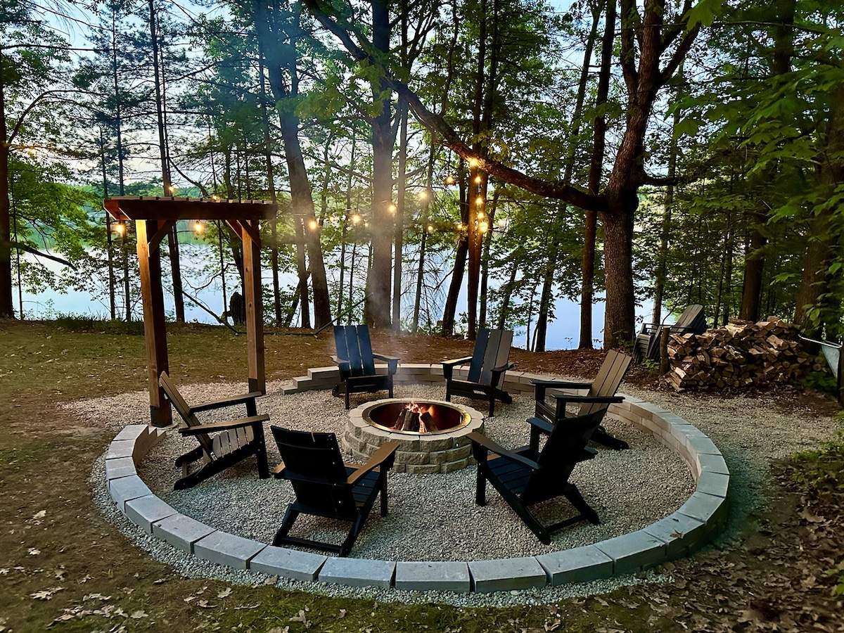 A stone fire pit is surrounded by several Adirondack chairs on a gravel surface, providing a cozy gathering space. Soft lighting hangs from above, and the backdrop features trees and a lake view, creating a serene setting for evening conversations.