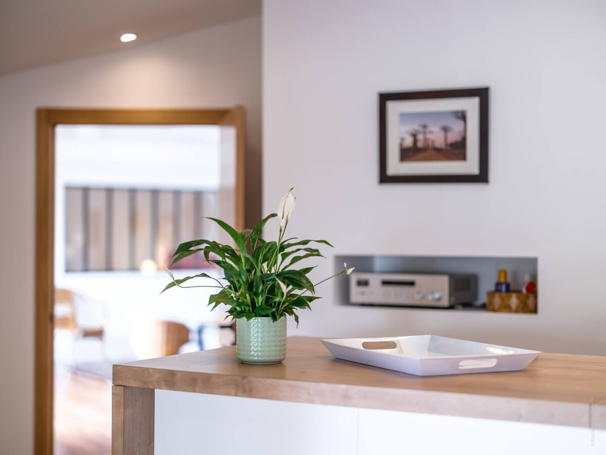 A bright and airy interior showcases a wooden countertop with a decorative white tray and a green potted plant. In the background, soft lighting and a hint of furniture can be seen, creating a welcoming atmosphere.