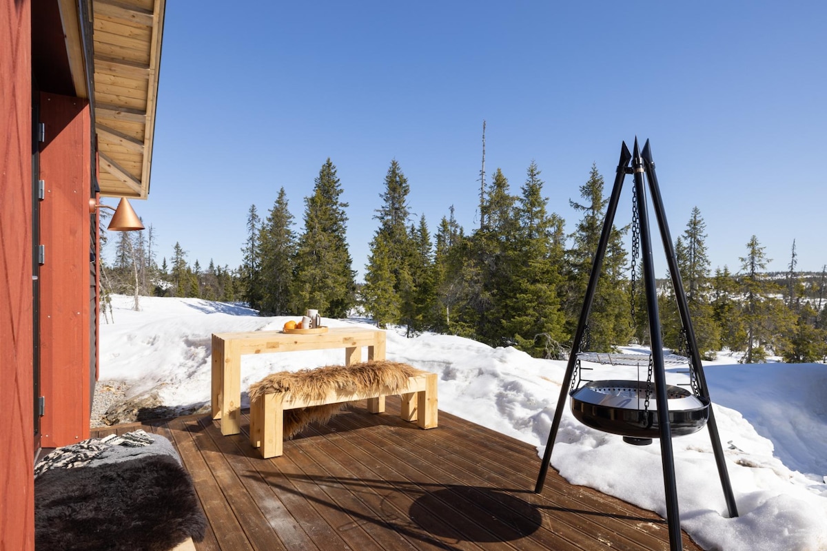 An outdoor seating area features a wooden table complemented by a fur throw, set against a backdrop of snow-covered terrain and evergreen trees. A modern fire pit is positioned nearby, with clear blue skies overhead creating a tranquil atmosphere.