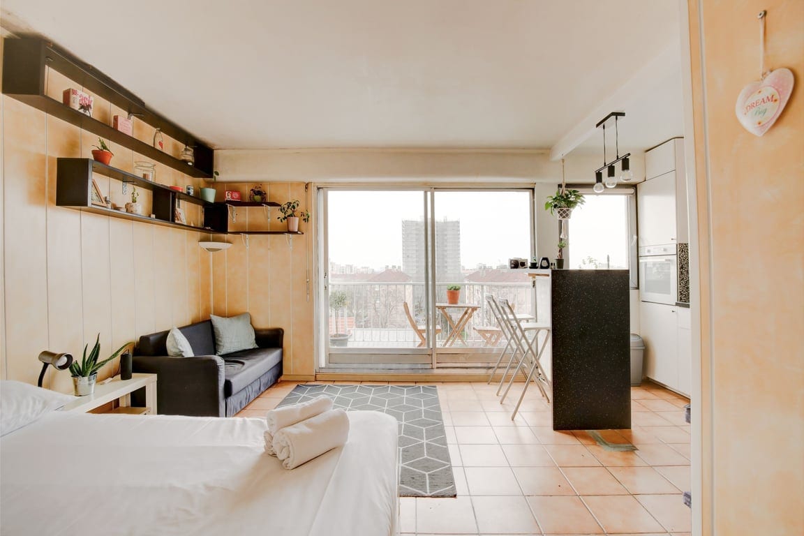 A spacious room features a comfortable sofa and a modern kitchenette with a black countertop. Large glass doors provide access to a balcony, and potted plants add a touch of greenery to the decor. The floor is tiled, creating a clean and inviting atmosphere.