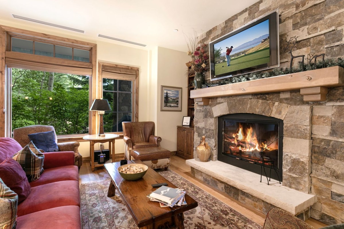 A cozy living area features a stone fireplace as the focal point, complemented by a comfortable couch and a leather armchair. A large window allows natural light and offers views of greenery outside. A flat-screen TV is mounted above the fireplace.