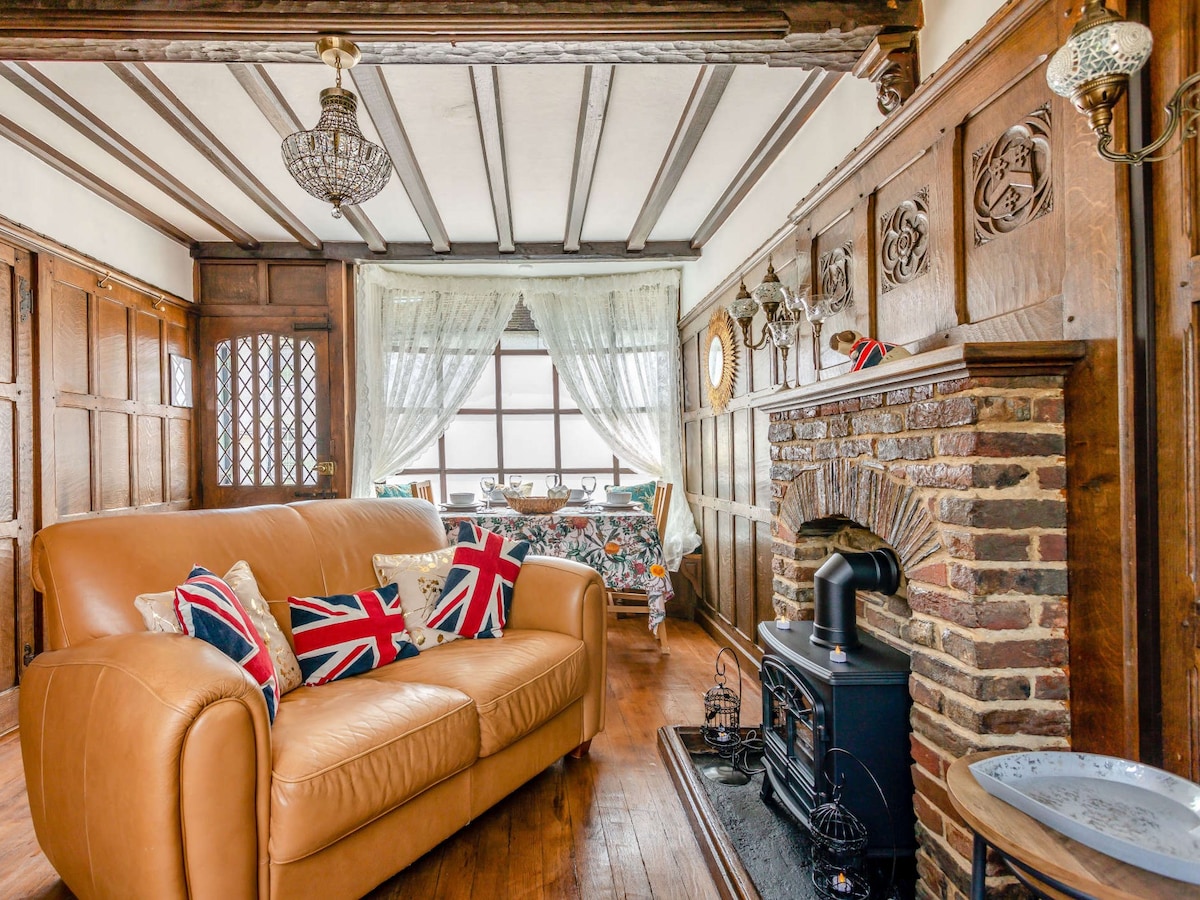 A warm lounge area features traditional exposed beams and wooden paneling. A leather sofa with decorative pillows is arranged around a wood burner effect stove. Natural light filters through large windows, enhancing the inviting atmosphere. A patterned tablecloth adds charm to the space.