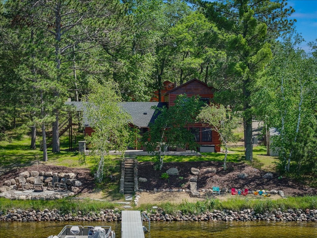 The cabin is nestled among tall trees, showcasing a two-story wooden structure with a welcoming porch. Stairs lead down to a landscaped area featuring natural stone and a peaceful waterfront view. A dock extends into the water, surrounded by lush greenery and colorful children's toys.