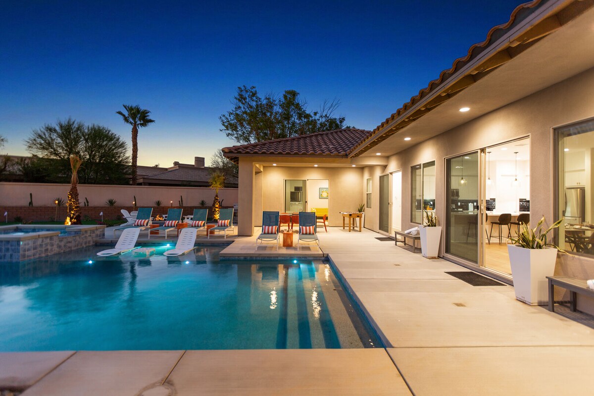 An inviting outdoor space features a private pool with a tanning shelf, surrounded by eight pool loungers and a seating area. The backdrop includes a charming home illuminated under a twilight sky, with palm trees visible in the distance.