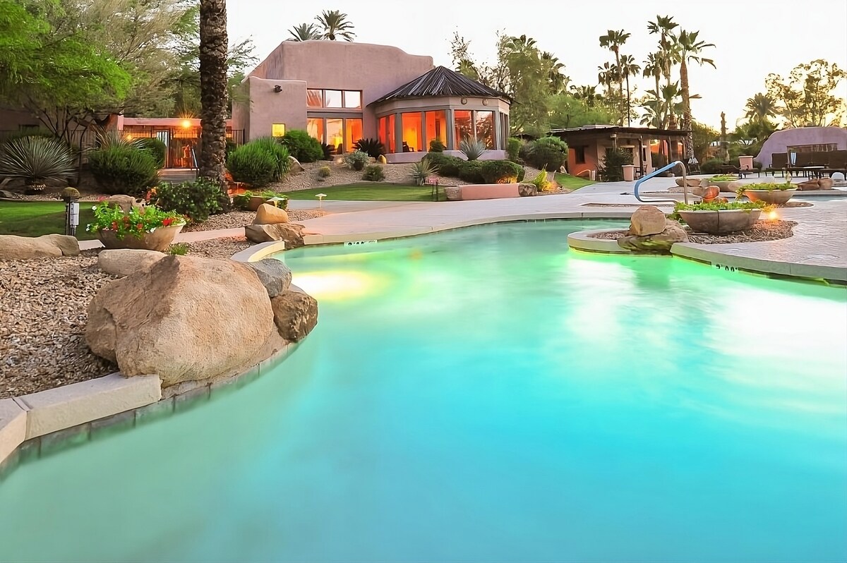 The outdoor pool area is highlighted, featuring a gentle curve with a smooth stone border. Soft turquoise water reflects ambient lighting. Surrounding landscaping includes well-kept gardens and palm trees, complementing the warm hues of the nearby building under a twilight sky.