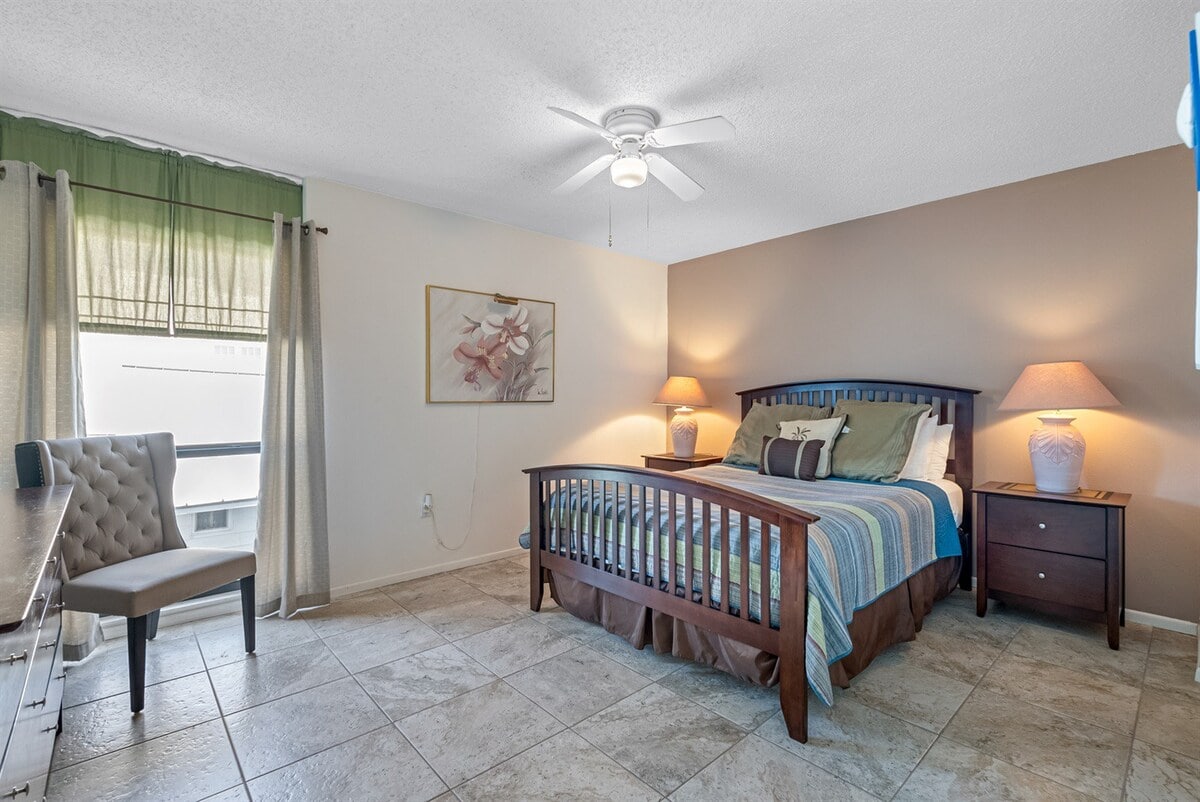 A spacious bedroom features a comfortable bed with a decorative blanket in soft colors. Two bedside tables with lamps are positioned beside the bed. A ceiling fan provides added comfort, while a window dressed with sheer curtains allows natural light to brighten the room.