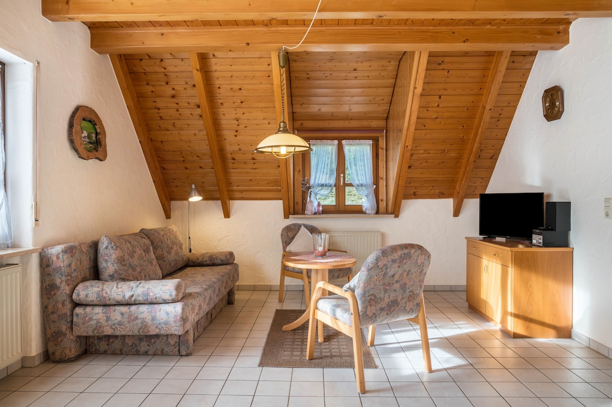 A cozy living area features a floral patterned sofa and armchair, complemented by a round dining table. Natural light is provided by a window with a decorative view, while wooden beams enhance the ceiling's warmth. A flat-screen TV is positioned for entertainment.