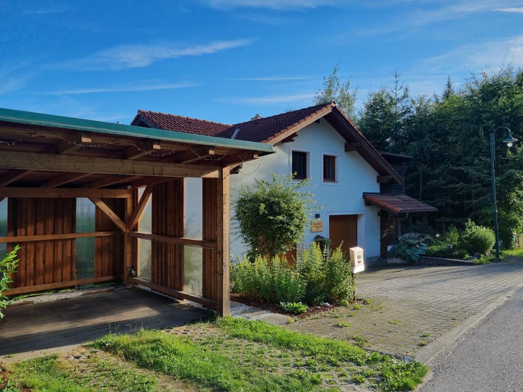 A rustic house is framed by lush greenery, featuring a sloped roof and wooden accents. A covered area for parking is visible on the side, and the entrance is marked by a welcoming sign. The surrounding nature creates a serene environment.