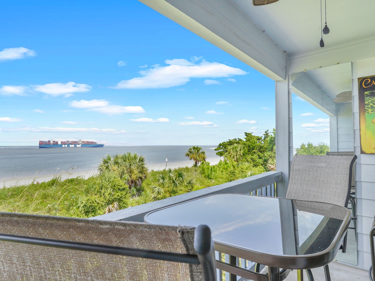 A balcony space features a glass table surrounded by comfortable chairs, poised for enjoying meals or relaxing. The view overlooks the expansive Atlantic Ocean, where a large cargo ship is visible against the horizon. Lush greenery encircles the area, enhancing the scene.