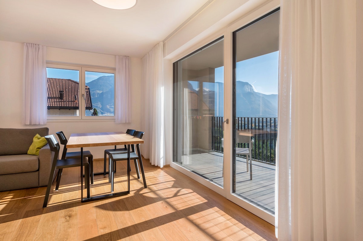 A bright living area features large glass doors that open to a private balcony, offering expansive views of the surrounding mountains. A wooden dining table with four chairs is situated in the space, complemented by a cozy sofa in a neutral tone. Natural light illuminates the room through the curtains.