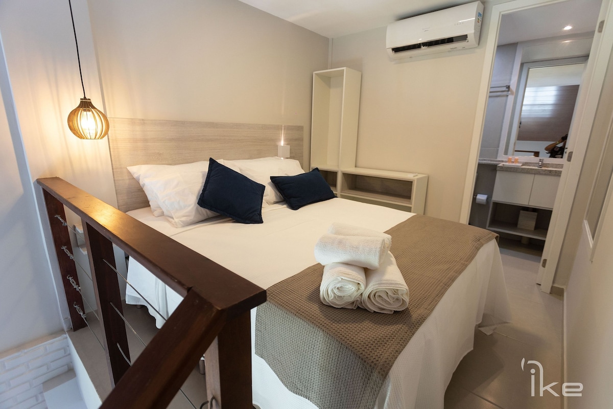 A compact bedroom features a spacious bed with neatly arranged pillows and a textured blanket. A warm pendant light highlights the space, while a modern air conditioning unit and a stylish cabinet provide comfort and functionality.