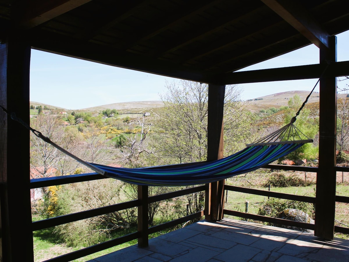 A colorful hammock is suspended from the wooden beams of a sheltered balcony, offering views of rolling hills and greenery. The natural light enhances the vibrant colors of the hammock, while the surrounding landscape includes trees and fields, creating a peaceful outdoor setting.