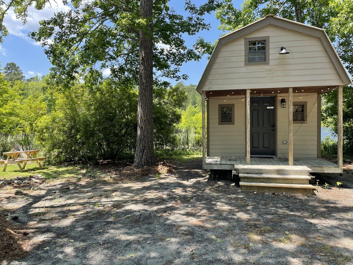 A rustic cabin is set among leafy trees, showcasing a welcoming porch and a dark front door. A picnic table is visible in the background, framed by greenery and gravel pathways. Natural light filters through the surrounding trees, enhancing the outdoor ambiance.