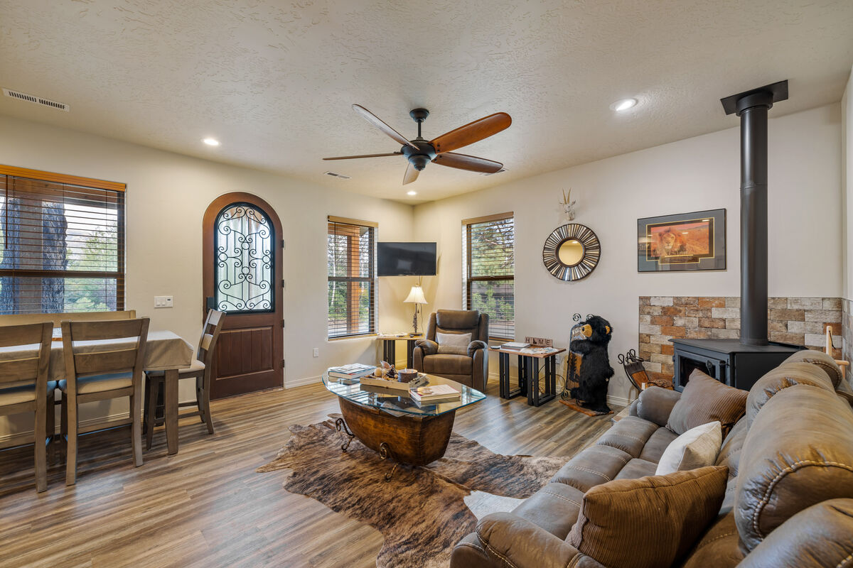 The living area is furnished with a comfortable sofa and armchairs, arranged around a unique wooden coffee table. Natural light enters through multiple windows, illuminating the space. A ceiling fan circulates air, and a wood-burning stove adds warmth to the inviting setting.