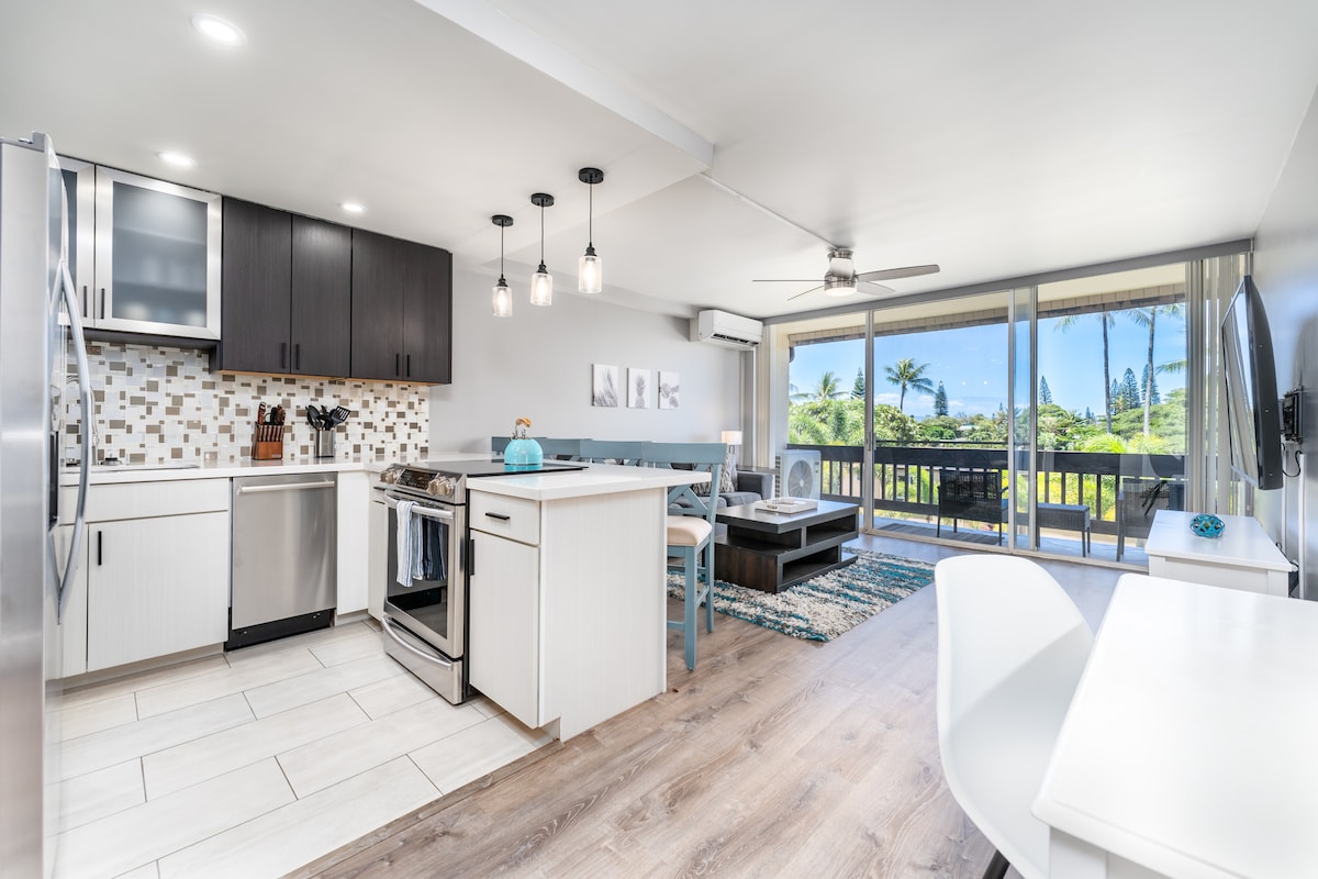 A modern kitchen is equipped with stainless steel appliances and offers a view into an open living area. Light wood cabinetry contrasts with dark upper cabinets. A spacious island doubles as a dining area, illuminated by pendant lights and facing large windows overlooking a lush outdoor space.