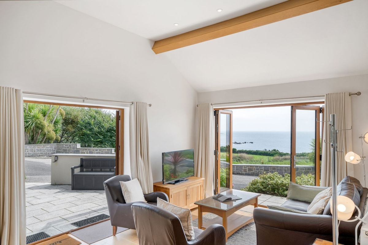 The open plan living area features large glass doors framing views of the sea. Two contemporary armchairs and a two-seater sofa face a wooden coffee table, with a TV cabinet nearby. Natural light fills the space, complemented by soft curtains.