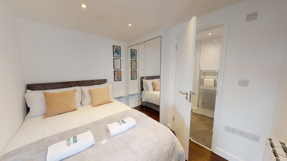 A well-appointed bedroom features a spacious bed covered in a light blanket. Two decorative pillows add subtle accents, while matching towels are placed neatly at the foot of the bed. A mirrored wardrobe and artwork create an inviting atmosphere, with an adjoining bathroom visible through an open door.