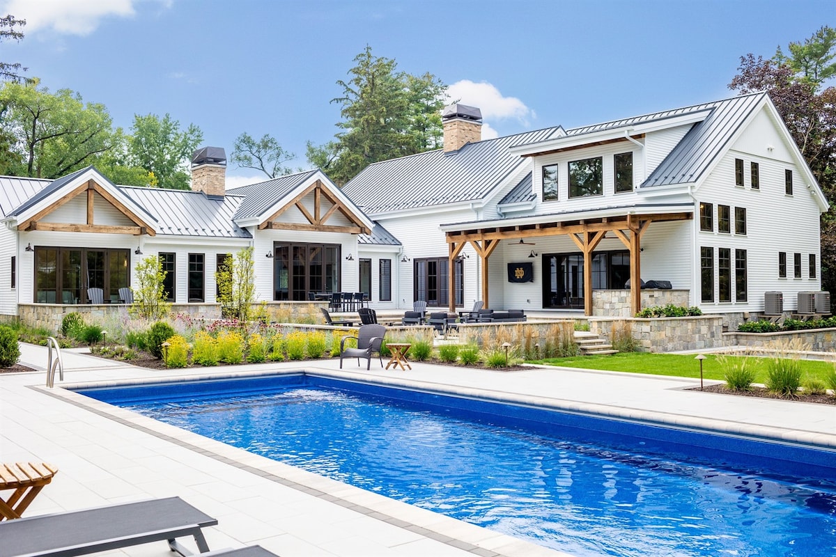 The spacious exterior features a large in-ground pool surrounded by paved decking. The modern farmhouse, with its white siding and metal roof, showcases multiple large windows and wooden accents, creating a harmonious blend of indoor and outdoor spaces. Lush landscaping enhances the serene atmosphere.