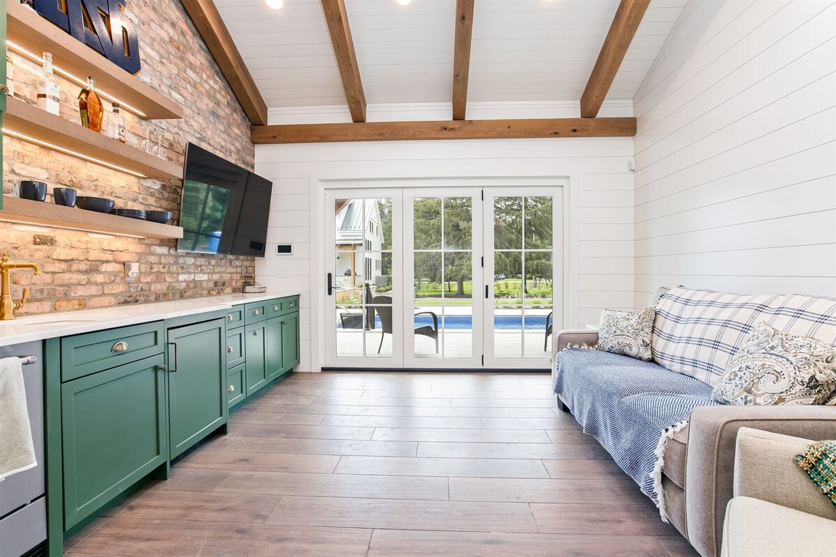 A cozy living area features a green kitchen cabinetry against a brick wall, accentuated by exposed wooden beams above. A comfortable couch is draped with a soft blanket, while large glass doors offer a view of the outdoor pool area.