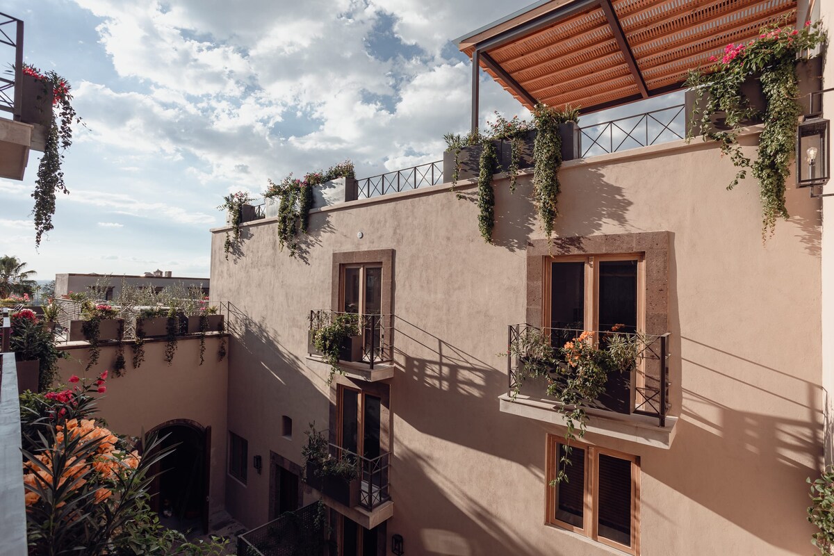 The exterior of the building is adorned with flowering plants cascading from balconies. Large windows offer glimpses into the light-filled spaces. A rooftop area is visible, shaded by a pergola, creating a serene retreat surrounded by greenery.