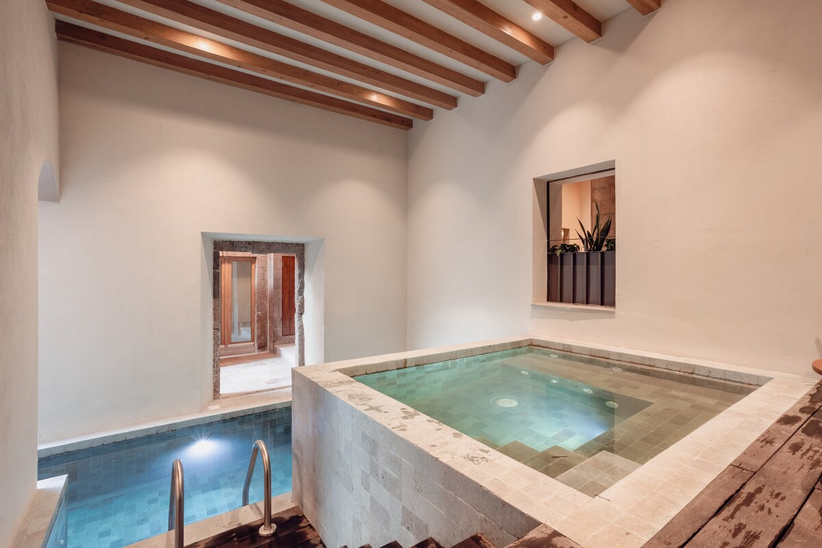 The spa-like area features a serene jacuzzi positioned next to a small pool, both surrounded by smooth stone tiles. Natural light enters through a nearby window, highlighting the neutral tones of the walls and wooden beams overhead.