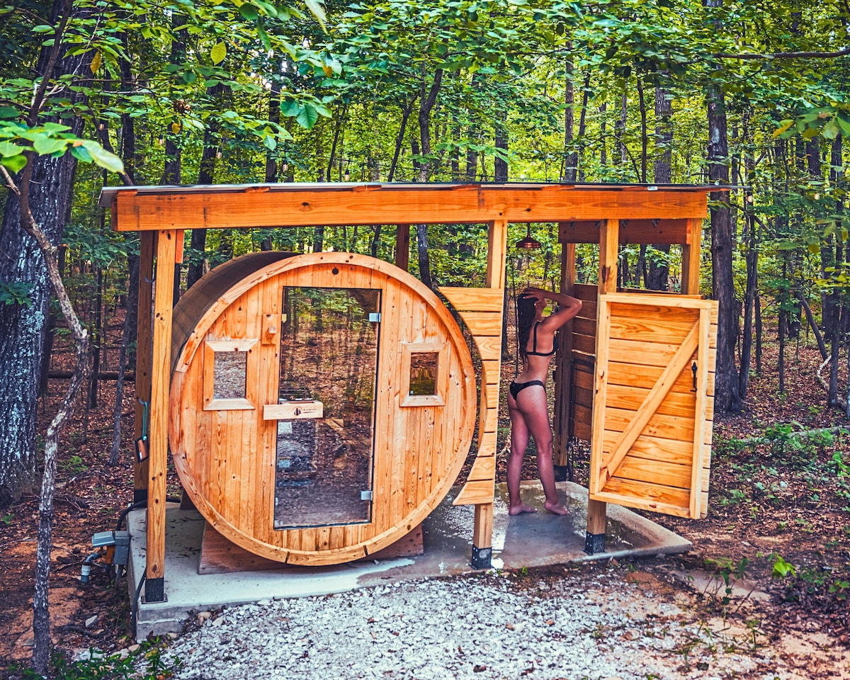 A wooden sauna structure is situated amidst a forest of lush green trees. The rounded design features a glass door and a wooden entry door, providing easy access. Natural stone flooring supports the sauna, enhancing the peaceful, nature-focused atmosphere.