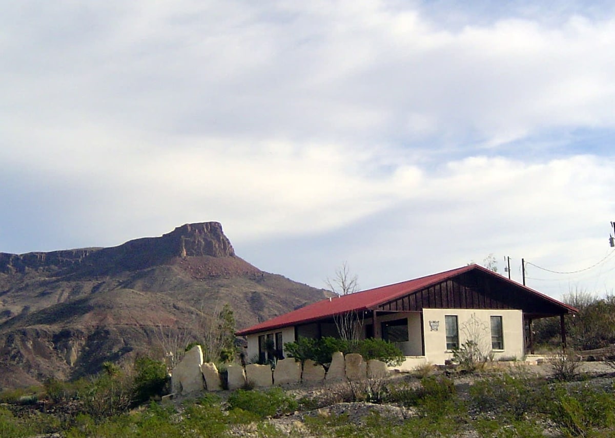 A modest house is set against a backdrop of rugged mountains, featuring a sloped red roof and a simple stone border. The structure is surrounded by sparse desert vegetation, with the sky above displaying soft, rolling clouds.