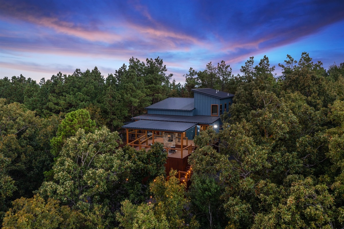 The cabin is nestled among towering pine and hardwood trees, showcasing a two-level design with large windows. The wrap-around deck is visible, providing ample outdoor space for relaxation. A serene color palette reflects the vibrant hues of the sunset against the sky.