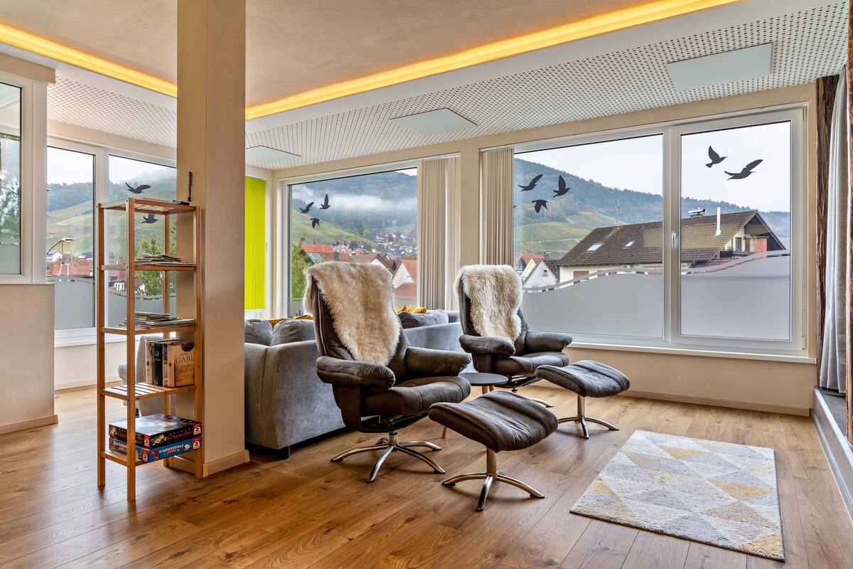 A bright living area showcases large windows offering views of the surrounding landscape. Two luxurious chairs with soft coverings are complemented by a cozy area rug. A wooden bookshelf holds various games, enhancing the space's inviting atmosphere.
