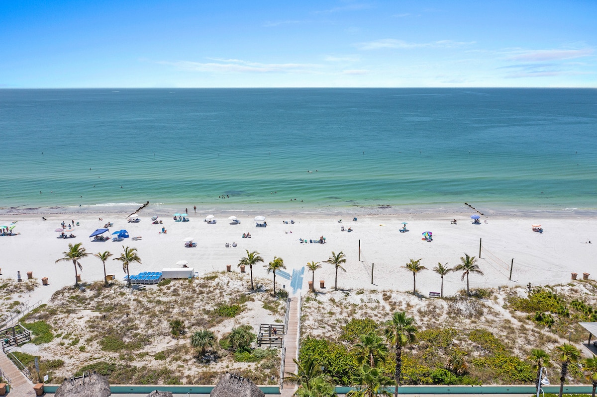 A scenic view of Madeira Beach is captured, showcasing soft white sands and gentle turquoise waves. Sunbathers and umbrellas can be seen on the beach, while palm trees line the foreground, creating a coastal atmosphere.