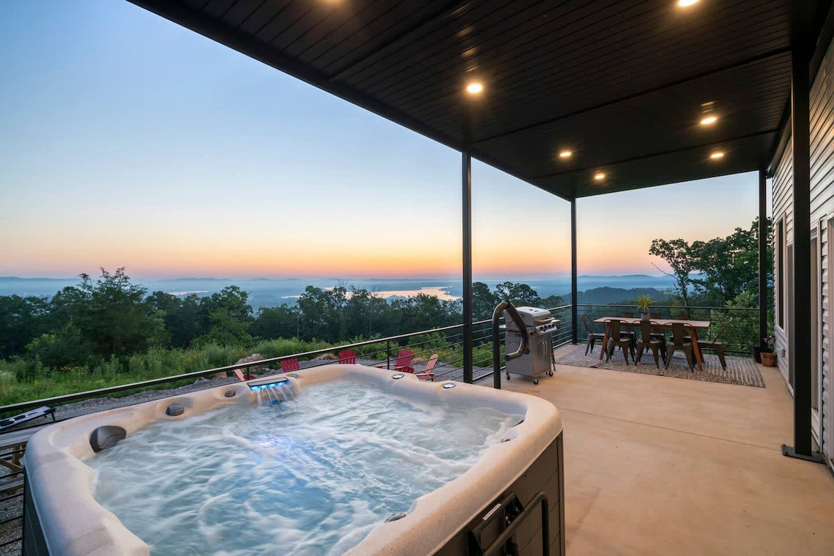 An outdoor patio area features a hot tub alongside a grill, with ample seating around a dining table. A scenic view of the landscape is visible beyond, complemented by a soft sunrise in the background.